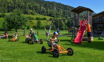 A playground with children riding go-karts. In the background, there is a slide and a swing, surrounded by green grass and trees.