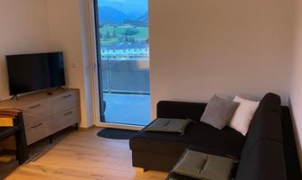 A modern living room with a dark sofa and a wooden table. The window offers a view of the landscape.