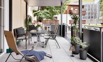 A modern terrace with a table, chairs, and plants. The view overlooks a green landscape and wooden buildings.