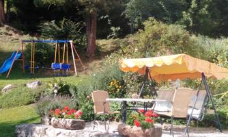 A beautiful garden with a swing and a slide. In the foreground, there is a table and chairs under a yellow parasol, surrounded by flowers.