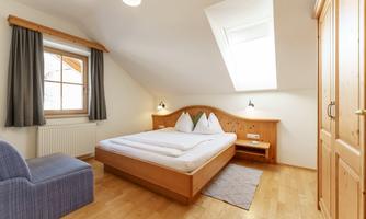 A bright bedroom with wooden floorboards and a double bed. The room has a window, a cozy seating area, and a wardrobe.
