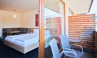 A modern room with a large window and a comfortable bed. Outside, there are two chairs and a small table on the balcony.
