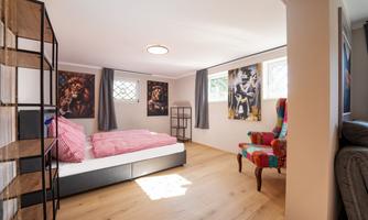 A modern bedroom with a double bed and a colorful armchair. The walls are adorned with artistic pictures, and the room has large windows.
