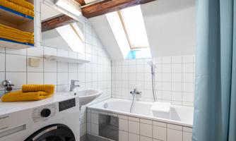 A bright bathroom with a window, a bathtub, and a washing machine. The walls are tiled and a shower cabin is present.