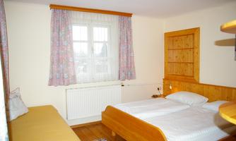 A cozy bedroom with a double bed and a sofa. The walls are light, with curtains and wooden shelves.