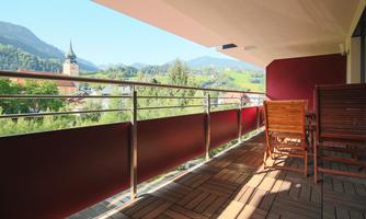 A spacious balcony with wooden chairs and a view of green hills and a church tower. The sun shines on the wood of the terrace.