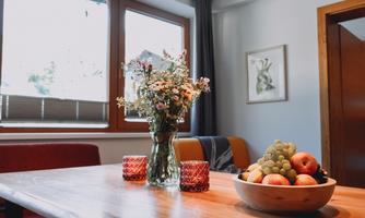 A beautiful table with a glass vase full of flowers and a bowl of fresh fruit. The windows offer a view of daylight and a cozy room.