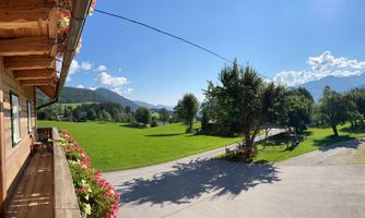 A beautiful landscape with green grass and trees under a clear blue sky. In the foreground, there is a balcony with flowers.