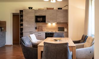 A modern dining room with a large wooden table and comfortable chairs. The kitchen in the background is functional and well-equipped.