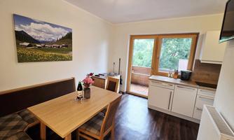 A cozy kitchen with a wooden table and chairs. Through the window, a green balcony with a view of nature can be seen.