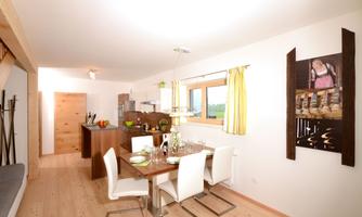 A modern dining room with a wooden table and white chairs. The kitchen in the background is well-equipped and offers a beautiful view outside.