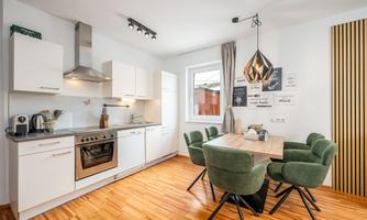 A modern kitchen with white cabinets and a wooden floor. A dining table with green chairs is in the center of the room.