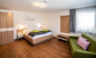A modern bedroom with a large bed and a cozy green sofa. The walls are light, and there is plenty of natural light coming through the window.
