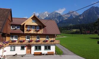 A charming house in the Alps with a garden and flower balconies. In the background, majestic mountains and a blue sky can be seen.