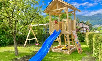 A beautiful playground with a wooden play castle and a blue slide. The surroundings are green with trees and a clear sky.