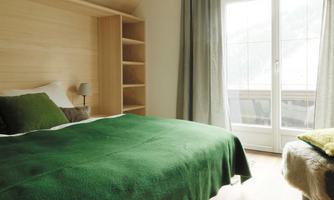A cozy bedroom with a large green bed and a window that leads to a balcony. The walls are made of light wood, and there is a bookshelf and curtains.