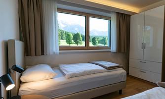 A modern bedroom with two beds and a large window. The view shows a green landscape with trees and mountains in the background.