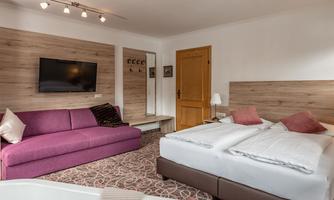 A modern hotel room with a double bed and a purple sofa. A television hangs on the wall and there is a small table.