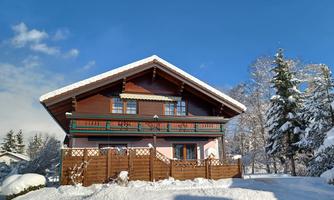A pretty chalet-style house surrounded by snow. The clear sky and the snow-covered trees create a picturesque winter landscape.