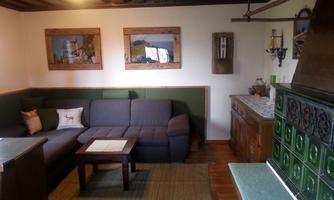 A cozy living room with a grey sofa and a wooden table. Pictures hang on the walls, and in the corner is a traditional stove.