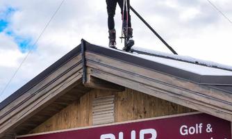 A man stands on the roof of a pub with skis in hand. In the background, there is a clear sky with a few clouds.