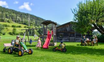 A playground scene with a playhouse, a slide, and children riding toy vehicles. In the background, green meadows and a farmhouse are visible.