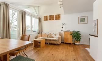 A bright living space with wooden flooring and large windows. There is a cozy seating area and a dining area.