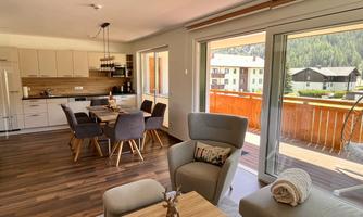 A modern living kitchen with a dining table and comfortable chairs. Large windows offer a view of the balcony and the surrounding mountains.