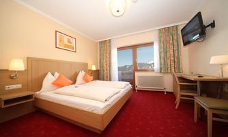 A cozy hotel room with two beds, a desk, and a television. Large windows let in plenty of daylight.