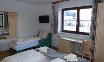 A cozy bedroom with two beds, a large window, and a small table. The walls are light, and there are wooden details as well as a television.