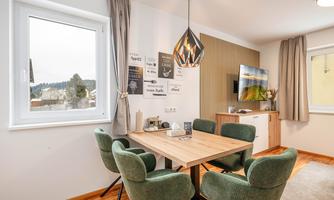 A cozy dining area with a wooden table and green chairs. Decorative items hang on the wall, and there is a television in the corner.