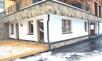 A modern building with stone and plaster facade. The surroundings are covered in snow and access is possible via a wooden terrace.