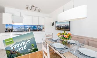 A modern kitchen with a large dining table and a colorful bouquet of flowers. In the foreground is a summer brochure offering vacation options.