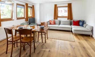 A bright living kitchen with a dining table and chairs. A cozy couch and large windows provide plenty of daylight.