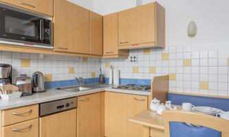 A modern kitchen with light wooden furniture and a gray countertop. There are appliances such as a microwave and a kettle, as well as a small dining area.