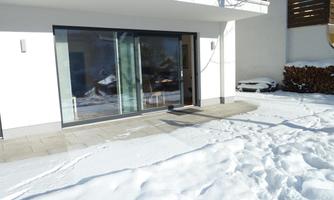 A modern residential building with large windows and a terrace door. The ground is covered with fresh snow.