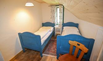 A cozy room with two blue beds and a wooden chair. The room has a wooden ceiling and a small window that lets in daylight.