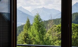 A view from an open window of green trees and mountainous landscapes. The clouds in the sky give the scene a relaxed atmosphere.
