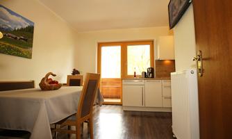 A friendly living room with a table, chairs, and a window front. The kitchen is modern and bright, with appliances and some fruits on the table.
