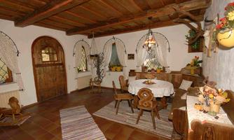 A cozy dining area with a round table and wooden chairs. The walls are adorned with rustic decorations, and large windows let in plenty of light.