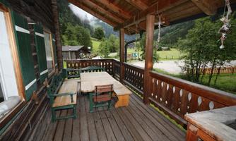 A wooden terrace with a table and chairs, surrounded by green hills. In the background, more buildings are visible.