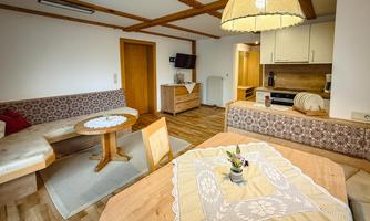 A cozy living space with a wooden ceiling and a round table. The decor is simple and inviting, with a small kitchen in the background.