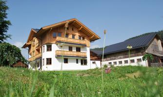 A modern wooden house with multiple floors and balconies. It is located in a green environment under a blue sky.