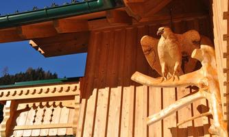 A wooden statue of an eagle is mounted on the wall of a traditional wooden house. In the background are trees and a blue sky.