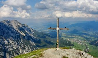 A cross on a summit with an impressive view of the surrounding mountains and the valley. The sky is partly cloudy and the landscape is green and hilly.
