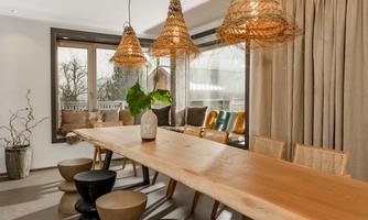 A modern dining room with a long wooden table and stylish, woven lamps. Large windows provide plenty of daylight and a view of nature.