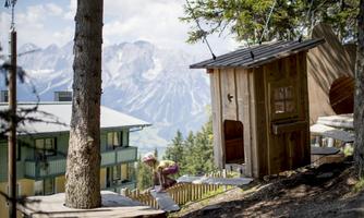 A playground in the mountains with a wooden cabin and play equipment. In the background, majestic mountains and a clear sky are visible.