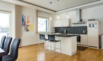 A modern kitchen with wooden flooring and bright furniture. There is a large work area with bar stools and plenty of natural light.