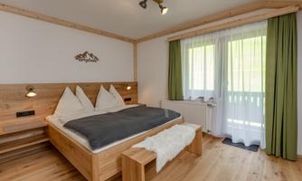 A cozy bedroom with a wooden bed and two pillows. Large windows provide plenty of daylight and a view of the balcony.