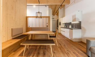 A modern dining room with a wooden farmhouse table and a bench. The walls are bright, and the kitchen is designed simply and functionally.